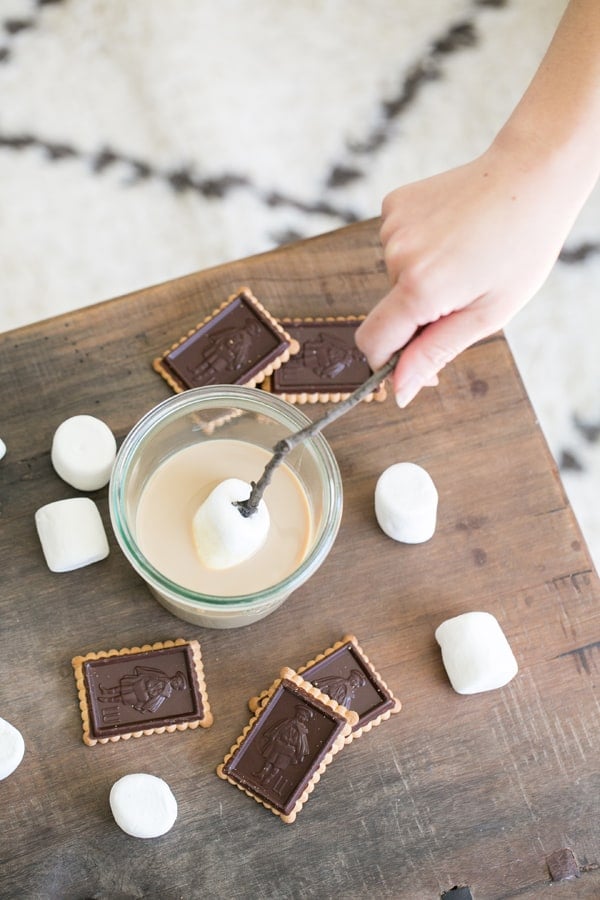 Hand dipping a marshmallow into a glass of Baileys Irish Cream with cookies.