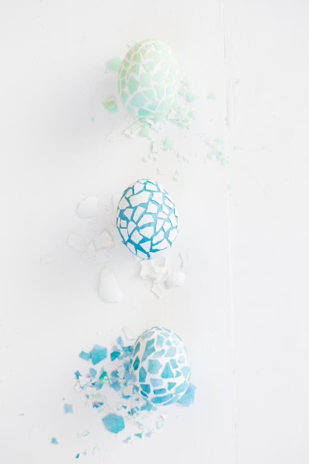 Green and blue Mosaic easter eggs in a row on a white table with cracked egg shells.