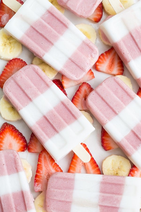 Striped strawberry and banana smoothie popsicles on a tray with sliced strawberries and bananas.