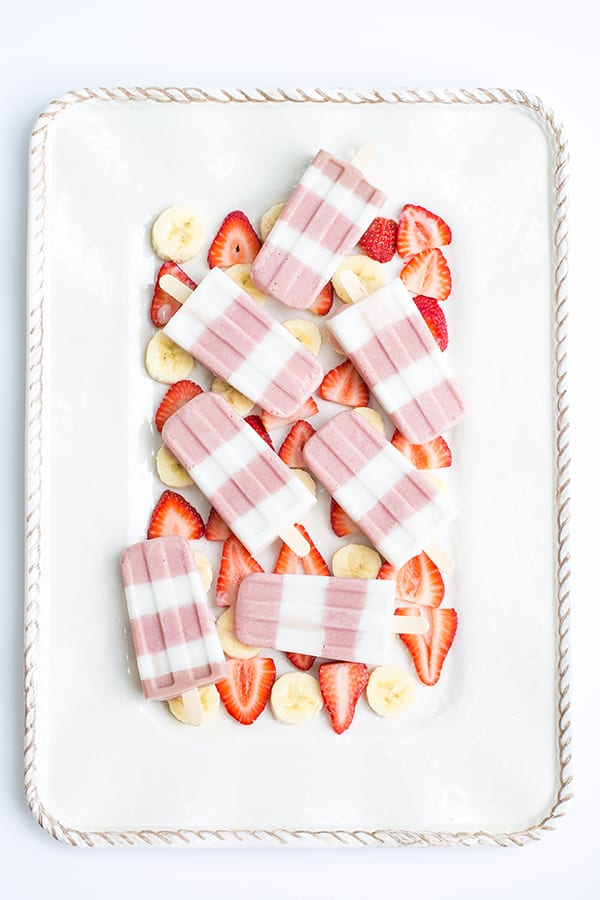 Striped strawberry and banana smoothie popsicles on a tray with sliced strawberries and bananas.