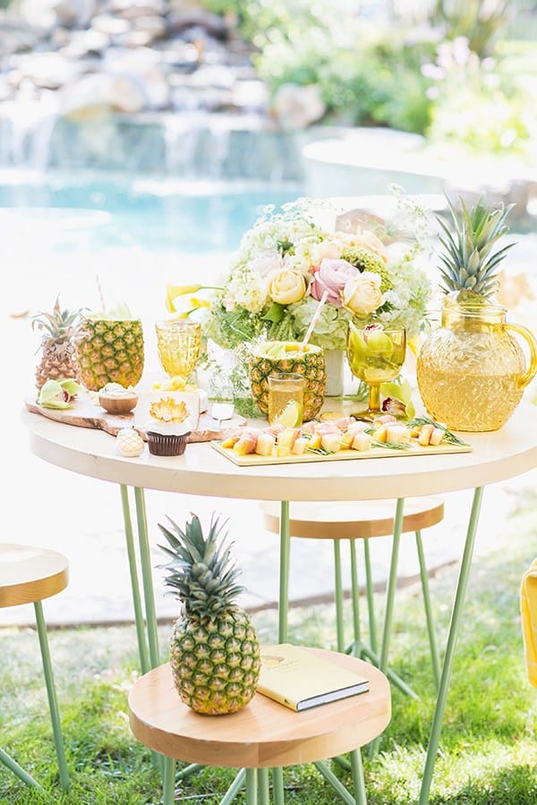 A pineapple party set up with food and drinks on a wooden table in front of a pool.