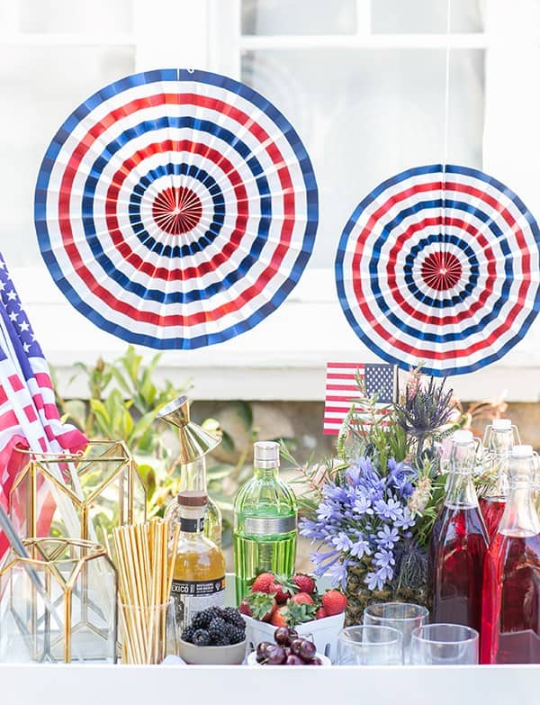 4th of July boozy snow cone stand with syrups, spirits, berries, paper fans and flowers.