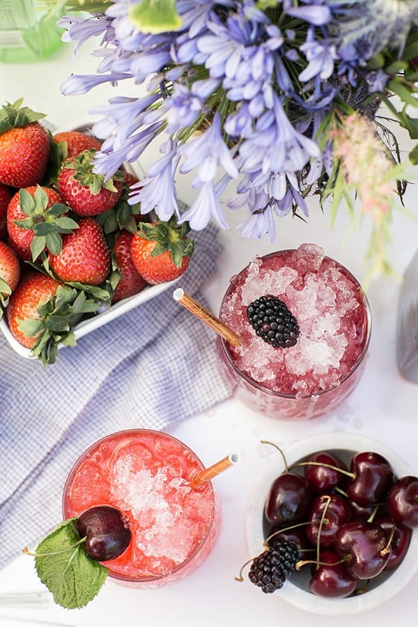 two boozy snow cones with berries and mint leaves and a bowl of cherries and strawberries 