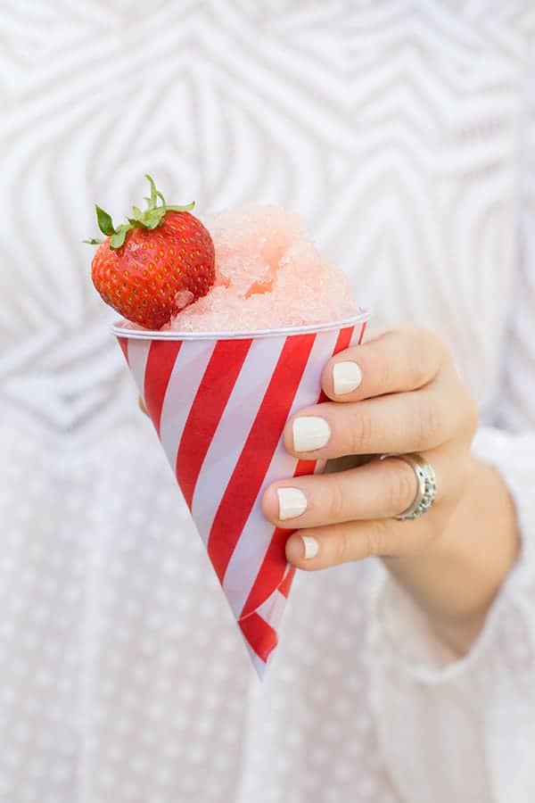 Holding a snow cone in a striped red and white cup with a whole strawberry on it. 