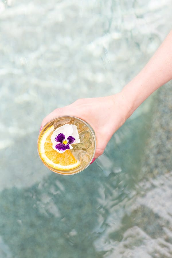 Hand holding a glass of pineapple party punch over a pool. It's garnished with an orange rind and edible flower. 