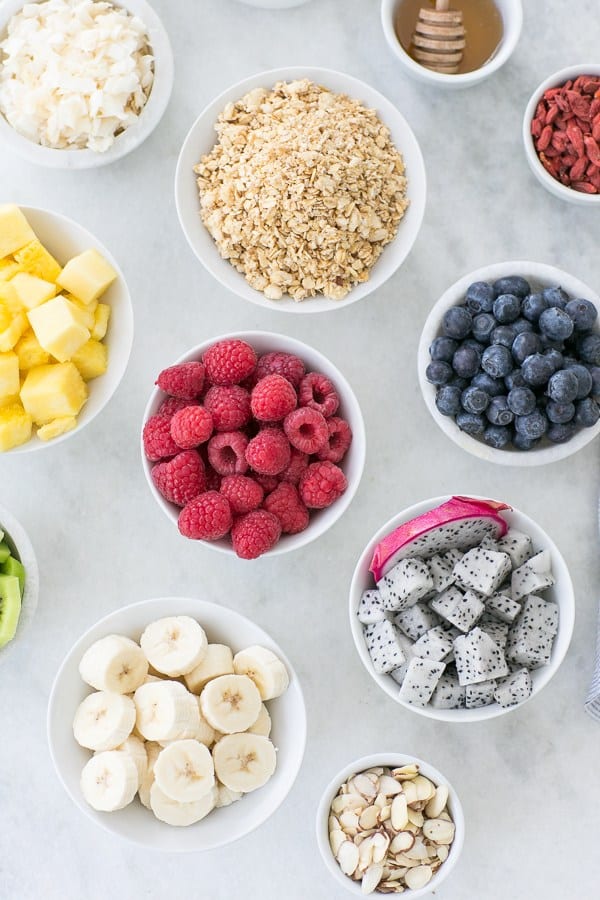 raspberries, blueberries, pineapple, bananas and coconut in small white bowls on a marble table.