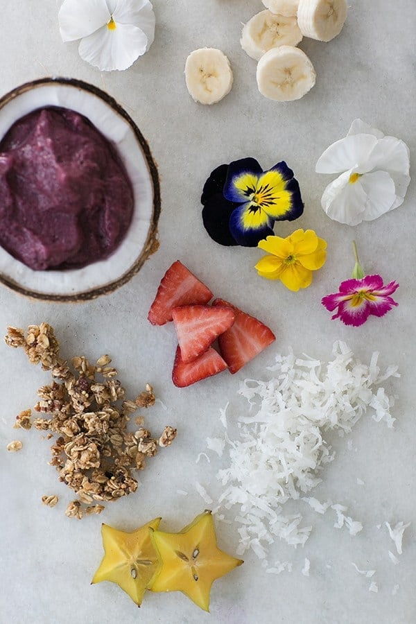 Sliced bananas, edible flowers, strawberries and granola on a marble table with an acai bowl.