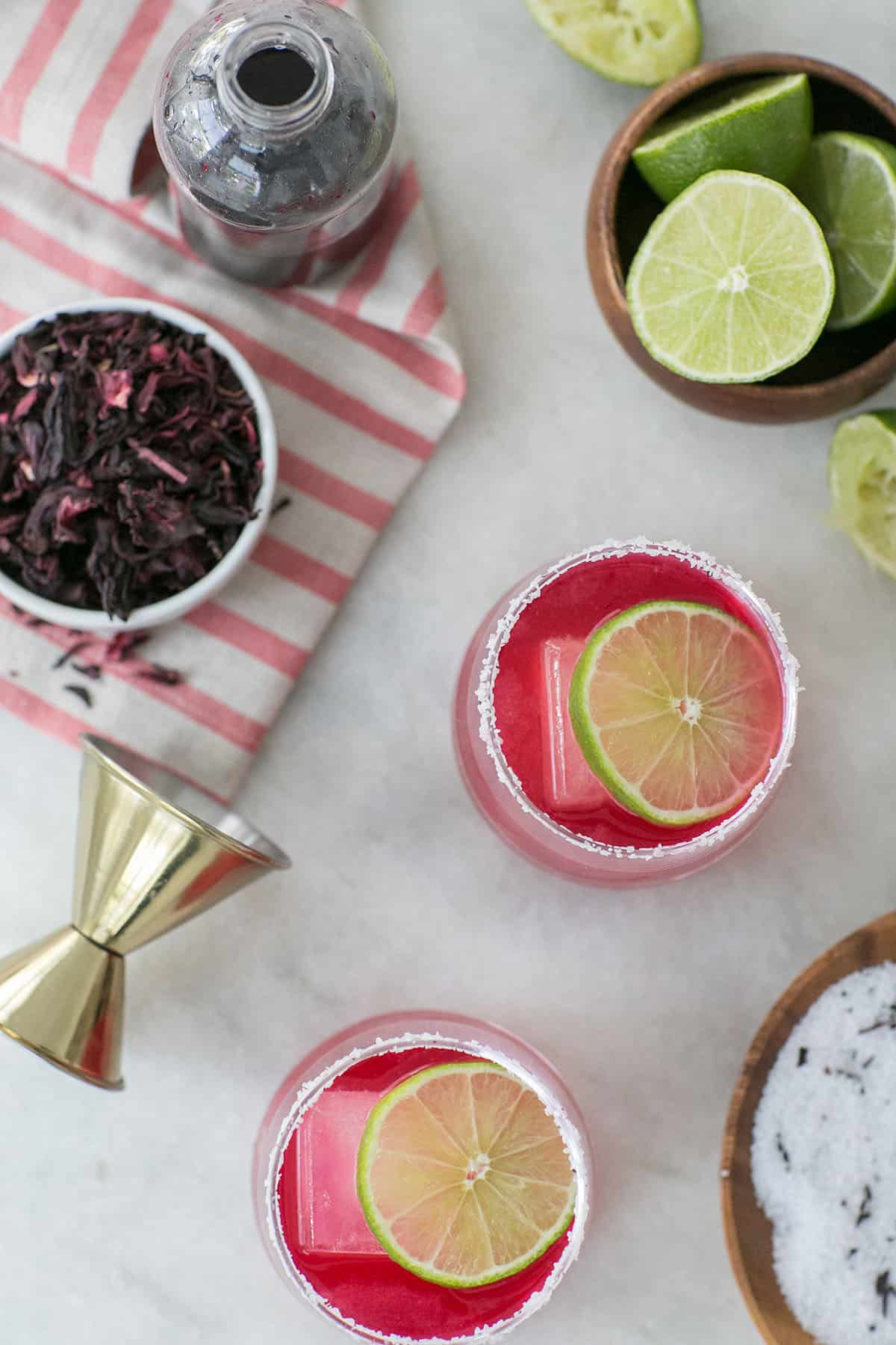 Vibrant pink hibiscus margarita on a table with limes and dried hibiscus flowers. 