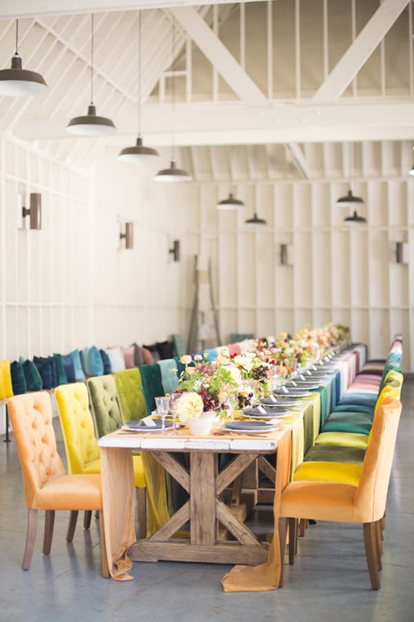 Long table inside a barn with colorful chairs and flowers. 