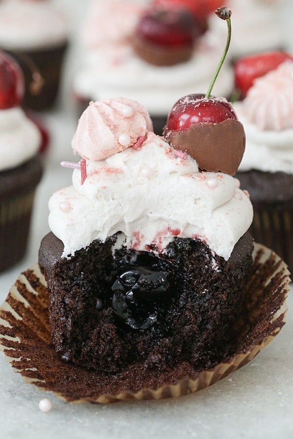 A chocolate cherry cupcake with a bite taken out of it, topped with whipped buttercream frosting and a chocolate cherry.