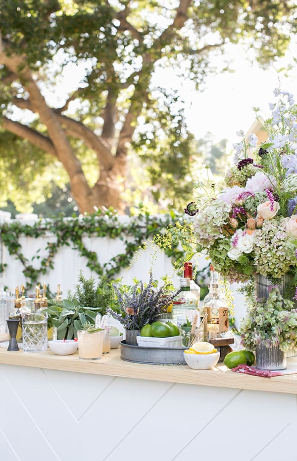 Whimsical garden cocktail bar setting with tons of flowers, herbs, limes, lemons and spirits.