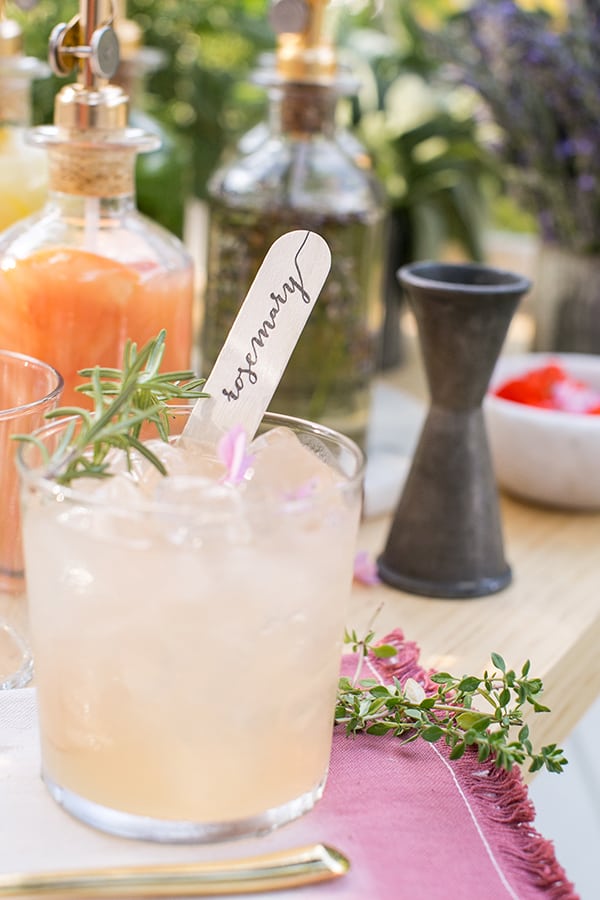 Pink cocktail with wooden stick and rosemary garnish.