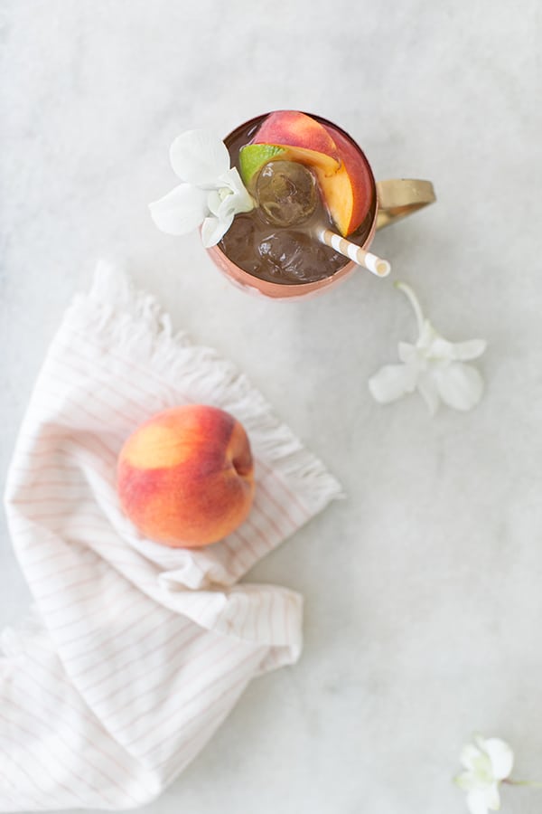 Peach Moscow Mule with a straw and ice and a peach on a napkin.