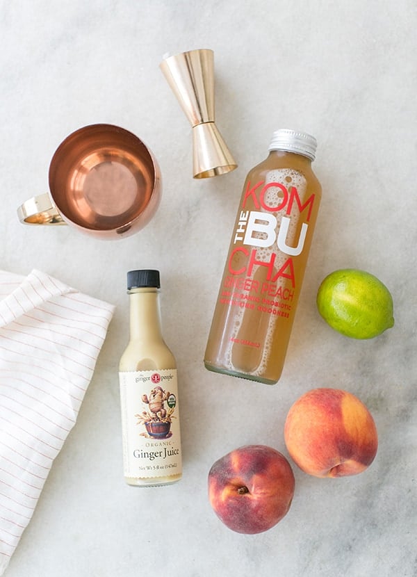 Kombucha, ginger syrup, copper mug, peaches and limes on a marble table.