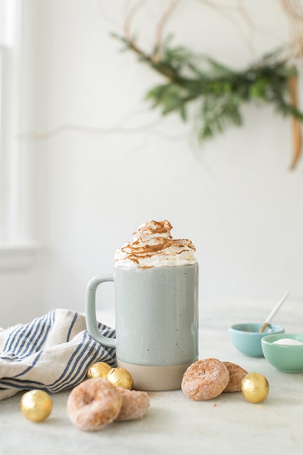 Blue ceramic mug on a table topped with whipped cream, cinnamon and mini doughnuts.