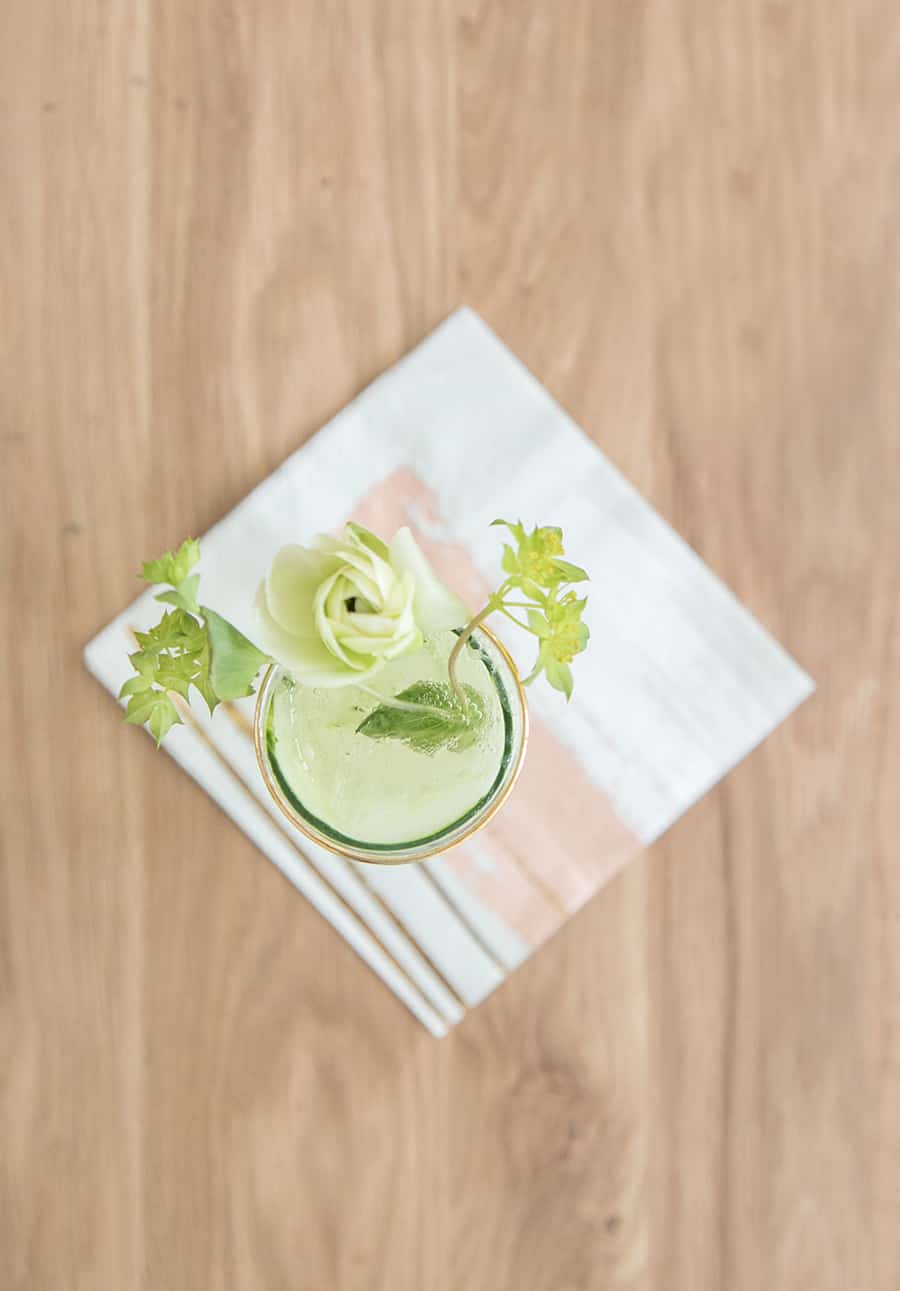 Cucumber and Kiwi Gimlet on a wooden table with flowers and a napkin.