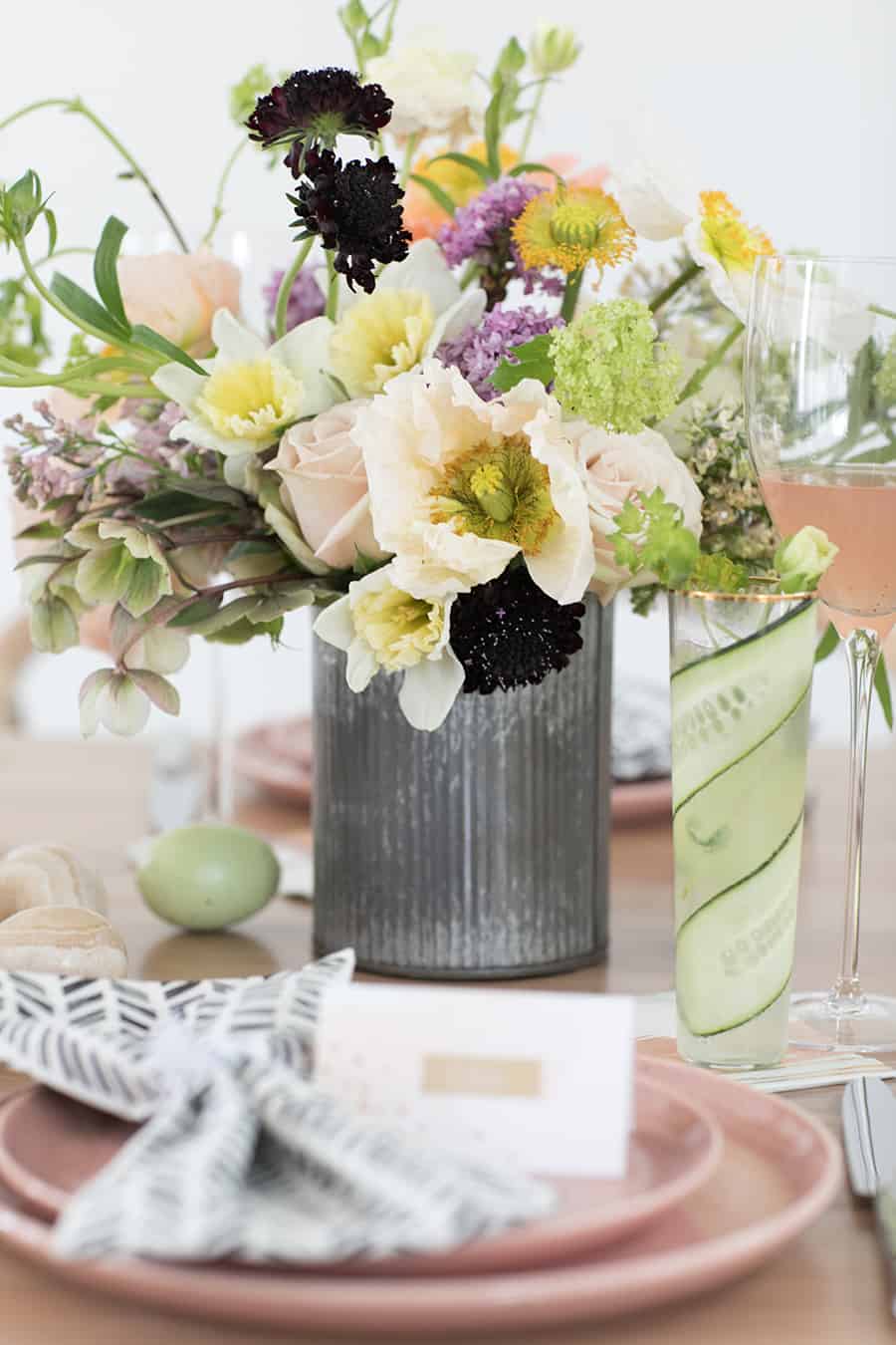 Colorful flowers in a galvanized vase on a table with a cucumber gimlet and Easter eggs.
