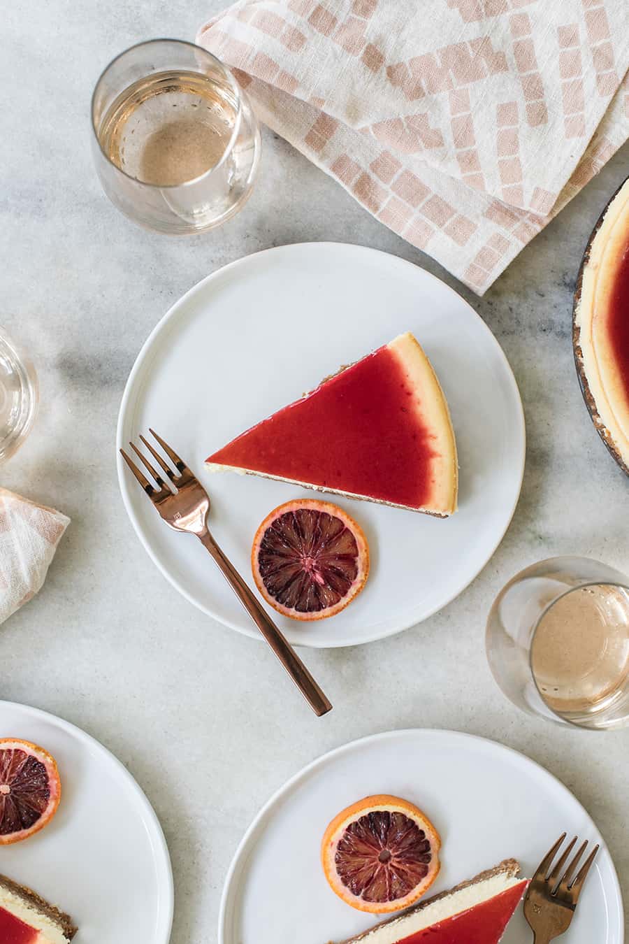 blood orange cheesecake on a white plate with a gold fork and slice of orange.