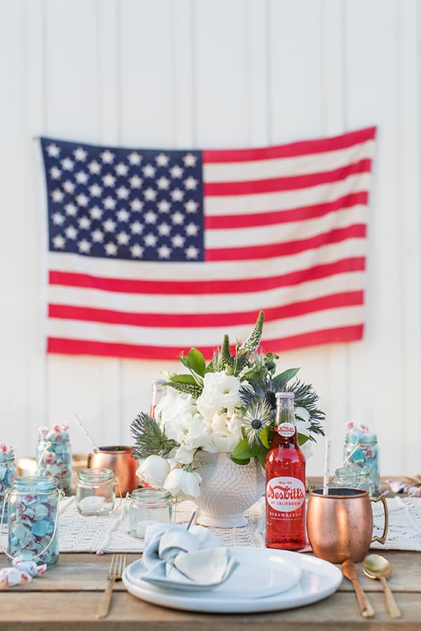 4th of July party with an American flag, vase of flowers, copper mugs, red sodas.