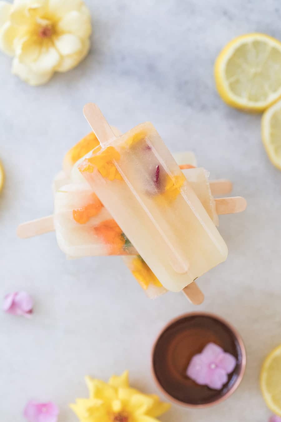 Three Ingredient Honey Lemonade Popsicles with edible flowers and lemons on a marble table.