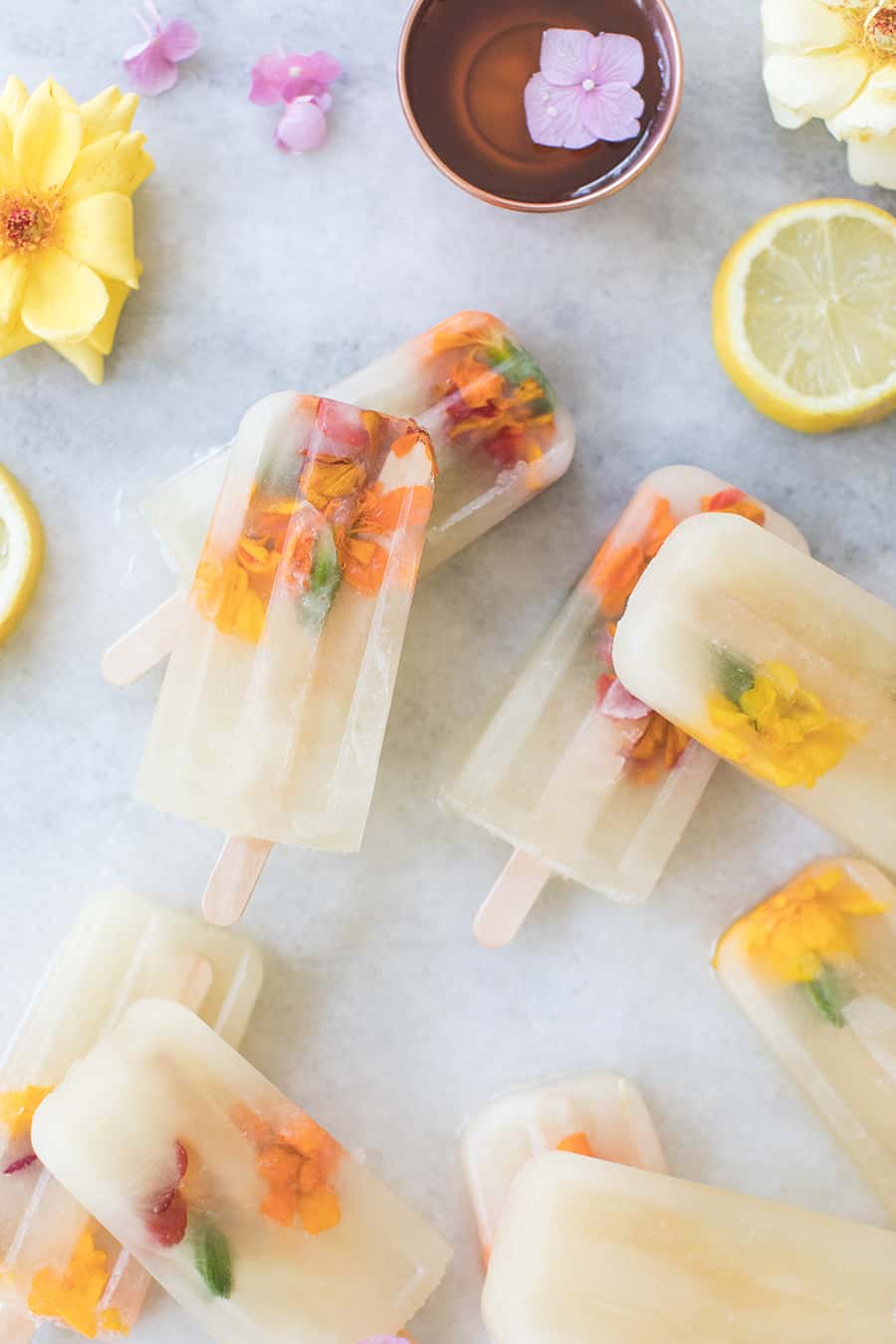 Tre Ingrediens Honning Lemonade Popsicles med spiselige blomster og sitroner på et marmor bord.