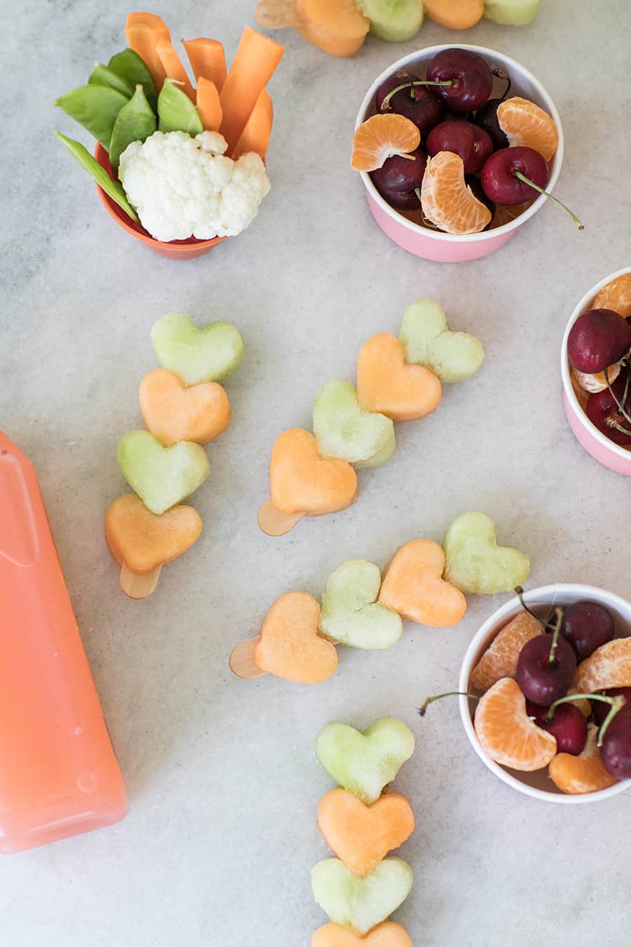 Heart fruit kabobs on a popsicle stick with a bowl of cherries and orange slices.