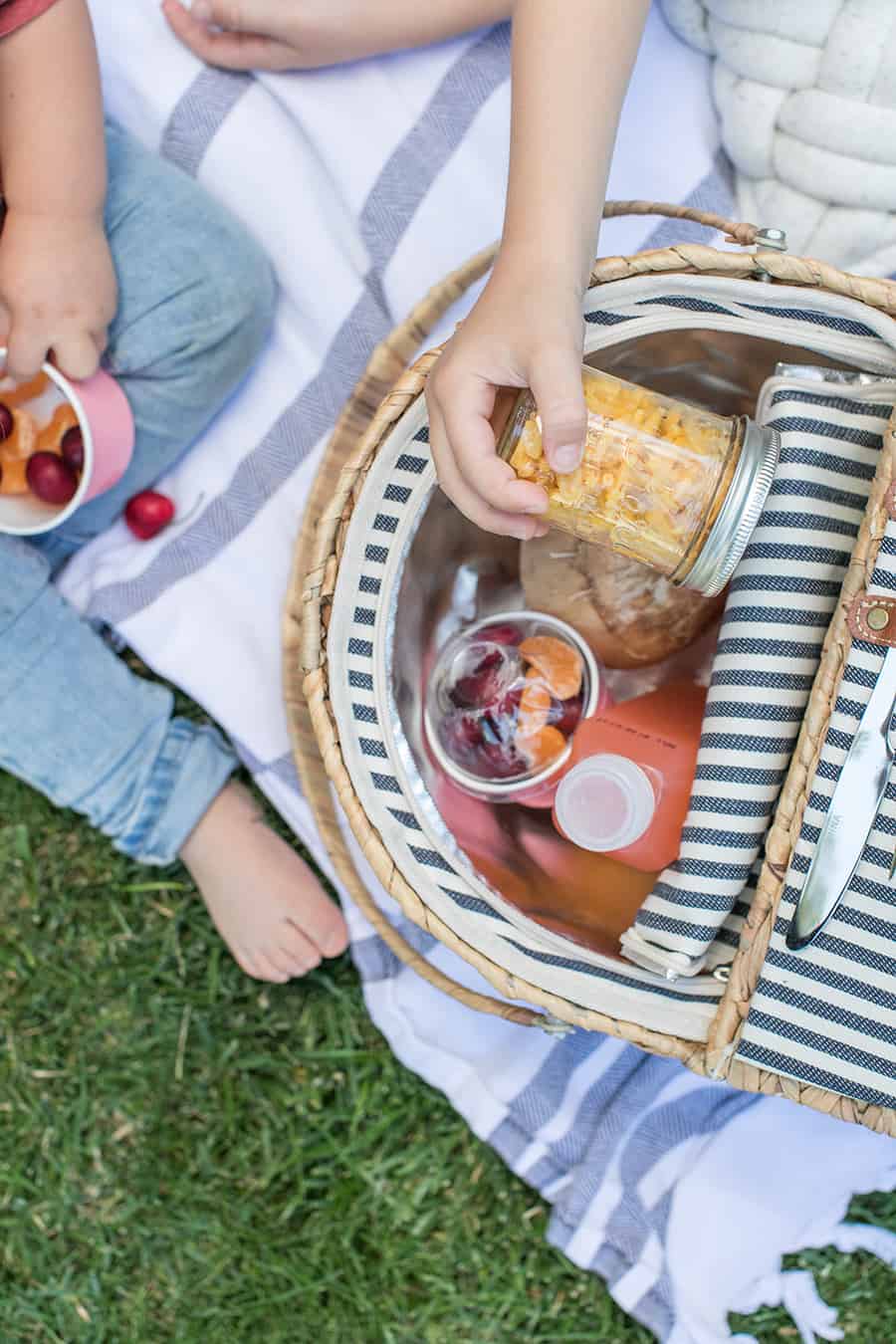 Food for kids in a picnic basket with a kids hand grabbing mac and cheese in a glass jar.