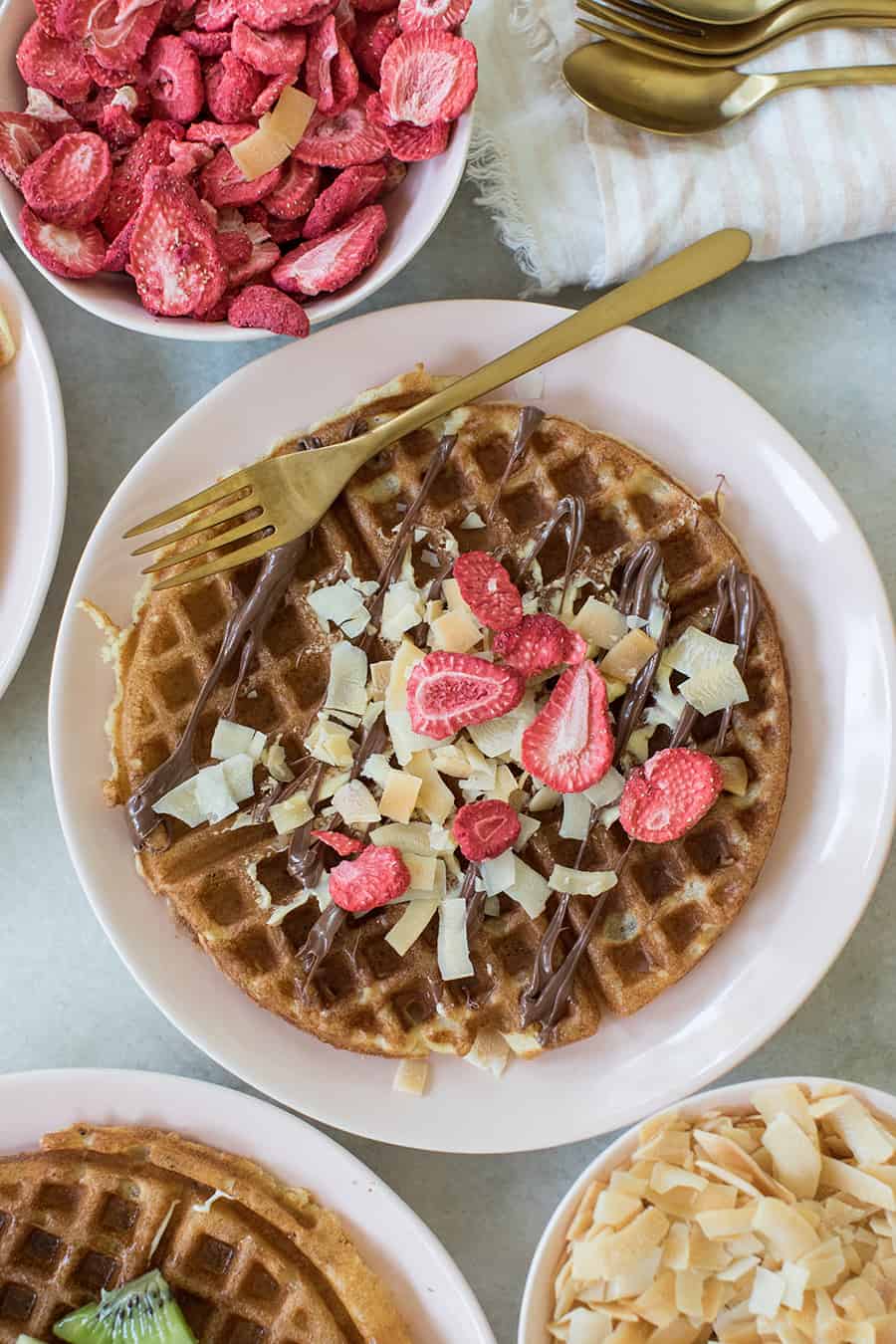 Waffle with Nutella, shredded coconut and freeze dried strawberries on a pink plate with a gold fork.