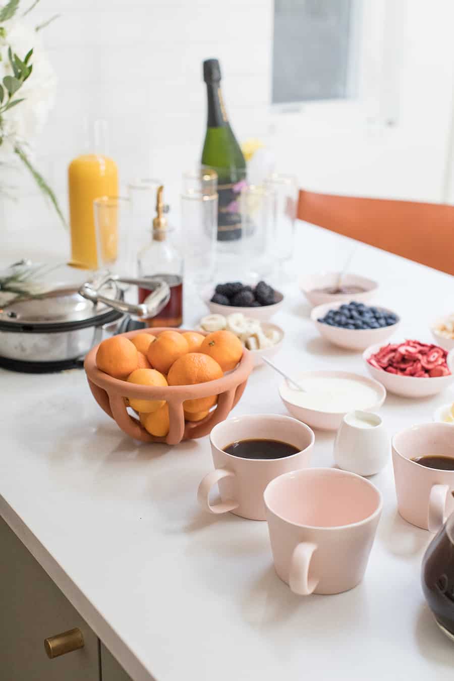 Waffle bar with pink coffee mugs, fruit, toppings and a waffle maker. 