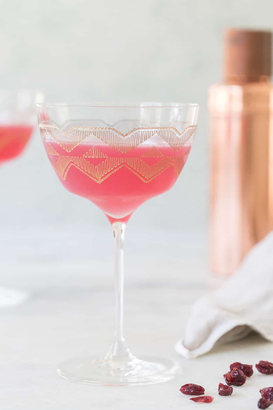 Pink cocktail in a coupe glass with gold etching and cranberries on the table.