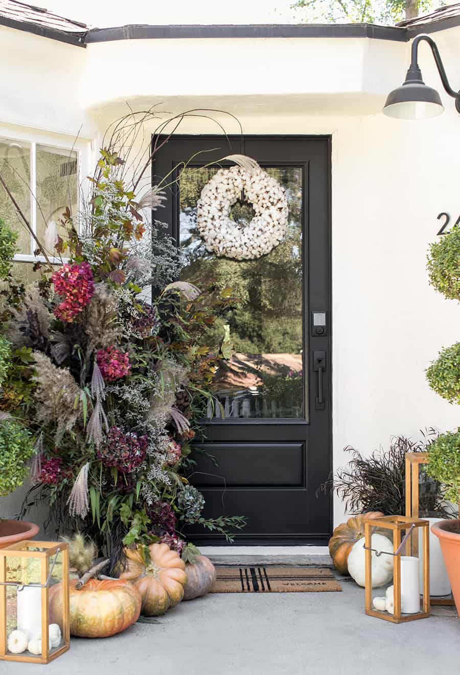 Fall front porch with black door and white wreath with flower arrangements and pumpkins. 