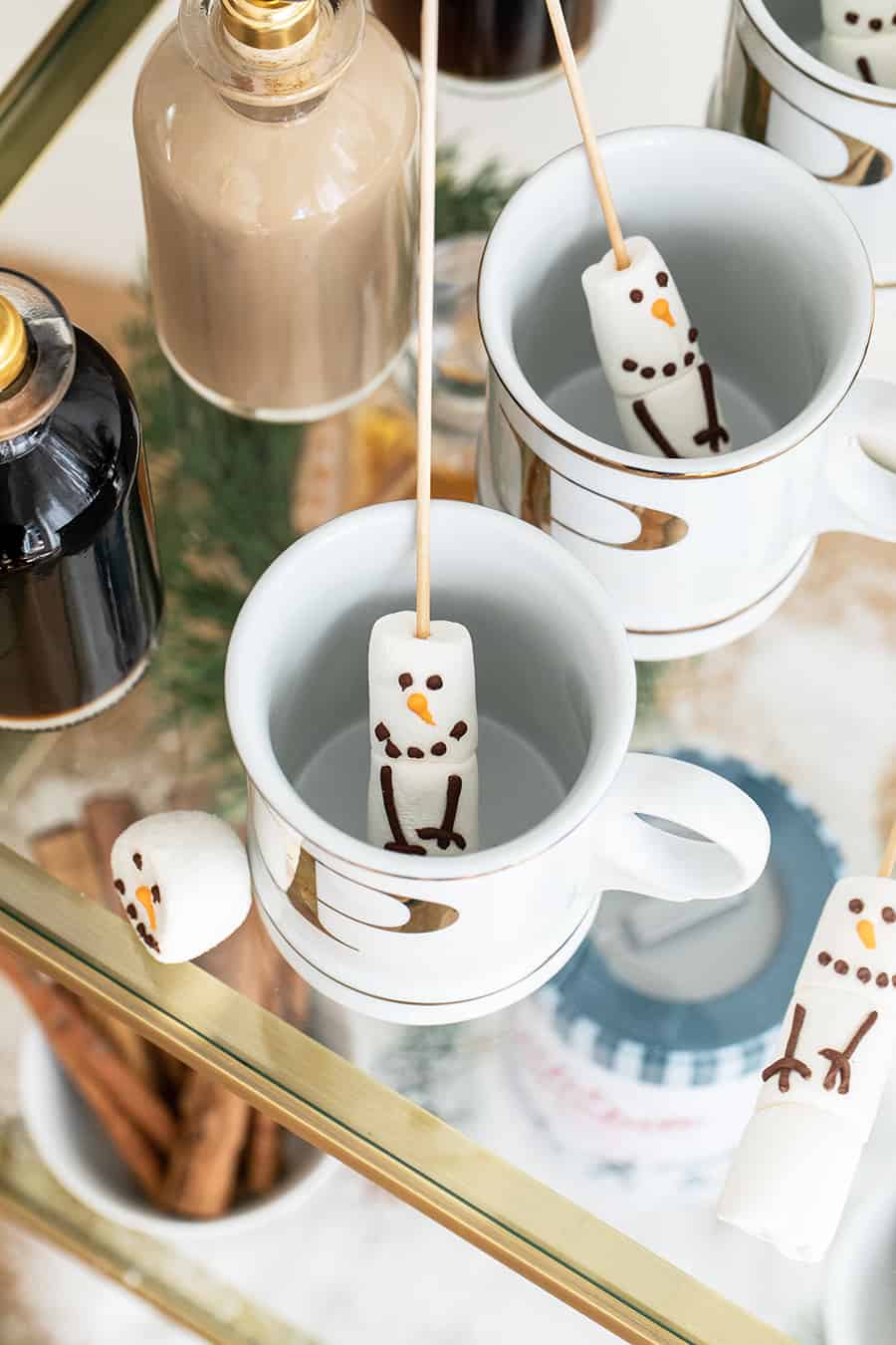 Snowman marshmallows on sticks going into a gold mug.