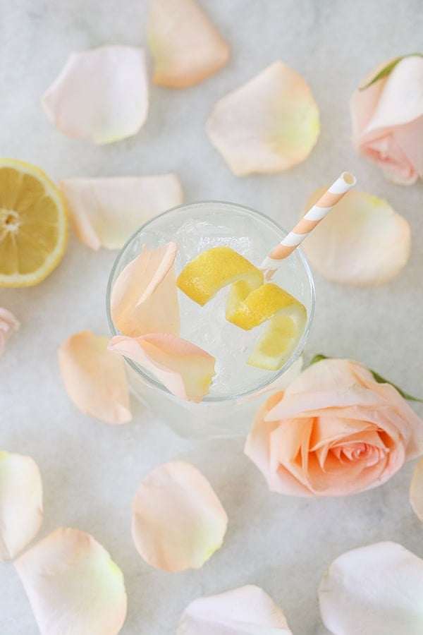 A cocktail with rose and lemon on a marble table with rose petals and a peach straw. 