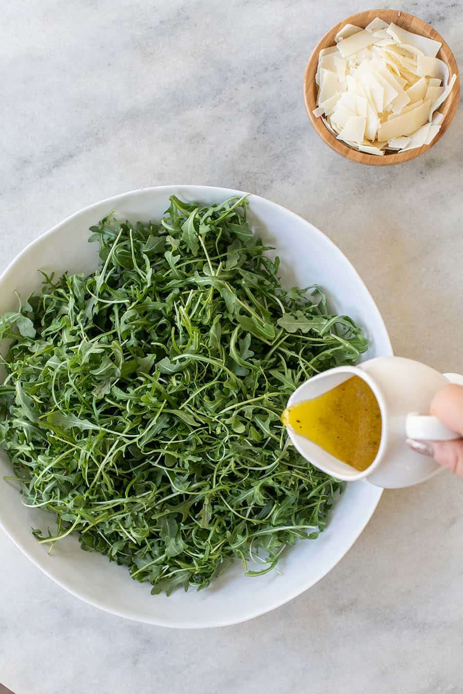pouring homemade dressing over arugula salad in a white bowl.