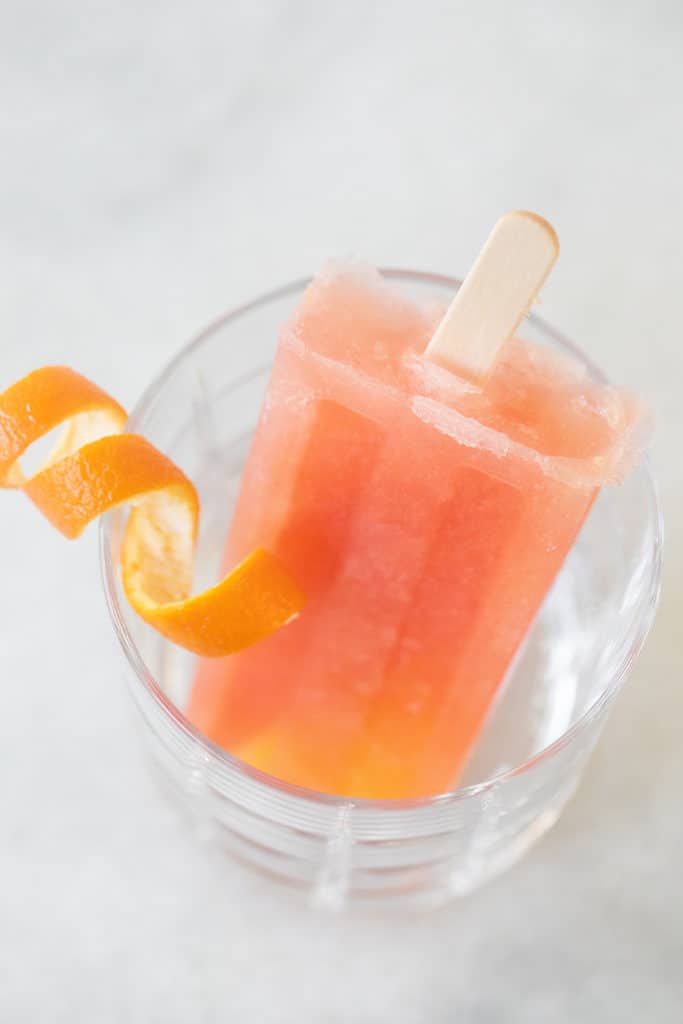 Pink fruit popsicles in a glass with an orange rind.