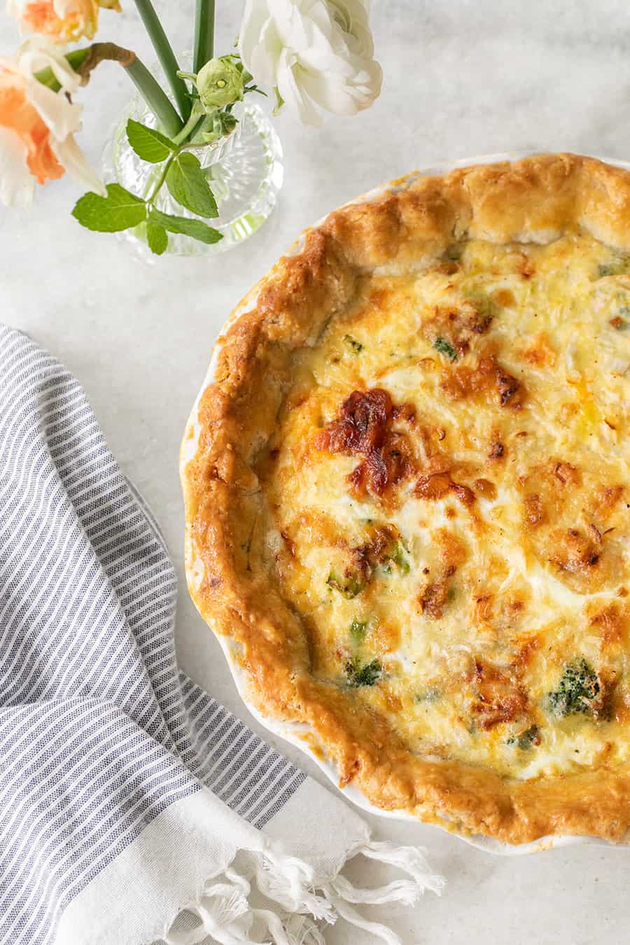 the most delicious quiche on a marble table with blue stripe towel and flowers.