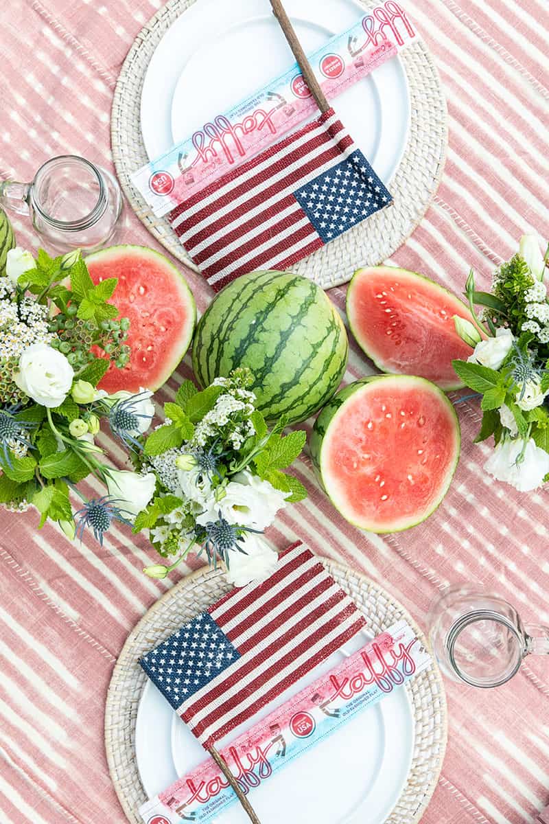 Table set for 4th of July party with watermelon down the middle, flags on table settings, mugs, pink table cloth.
