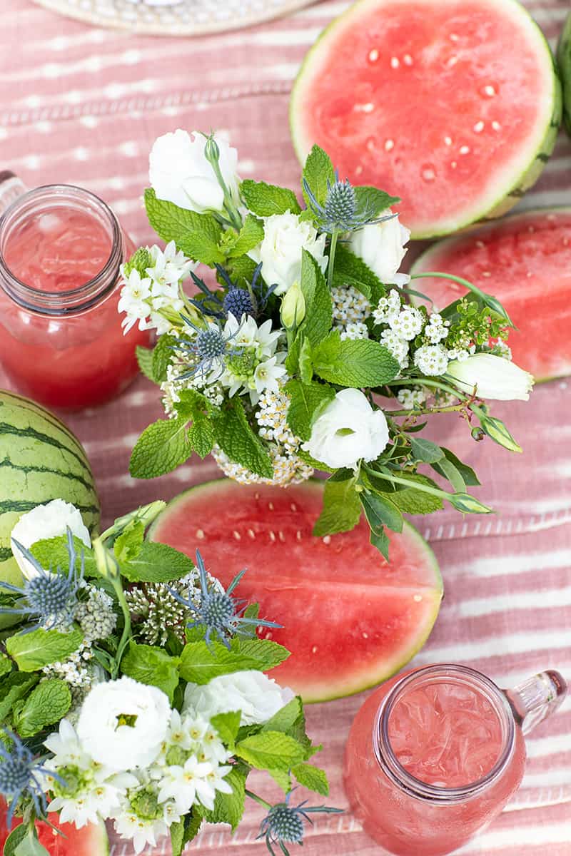 White and blue flower arrangement on a pink dyed table cloth with watermelons.