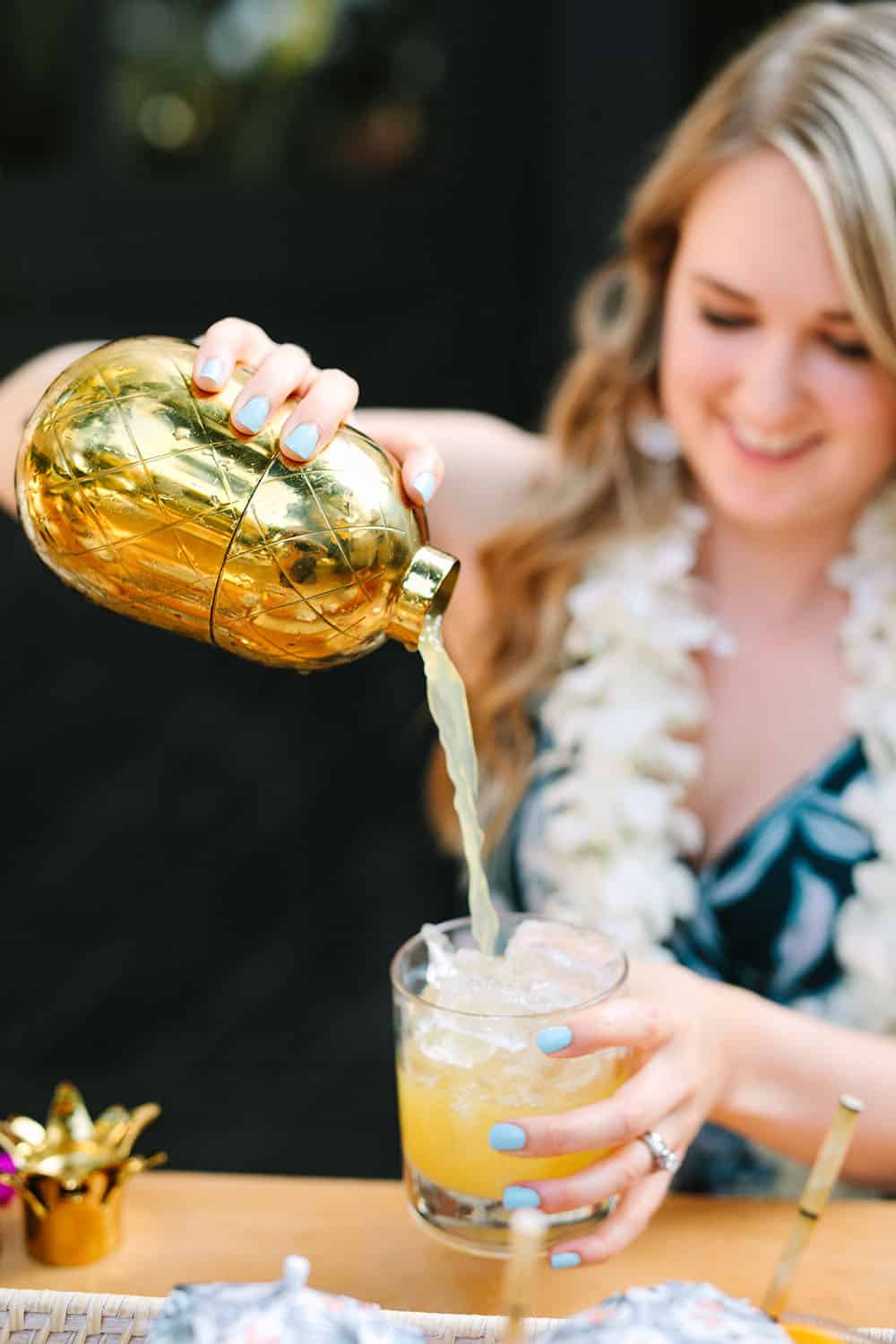 Girl pouring a tropical cocktail into a glass with a gold pineapple cocktail shaker.