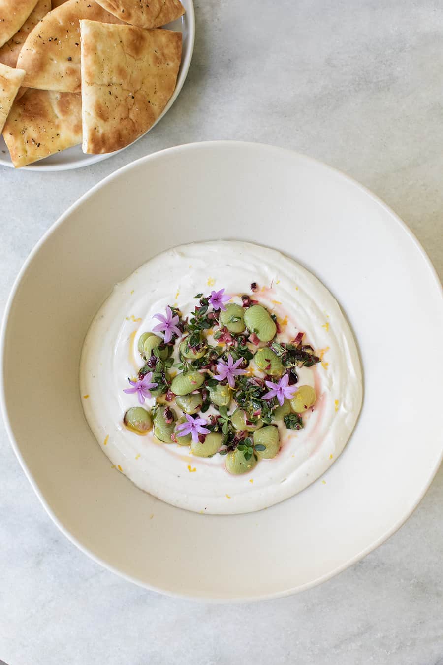 Yogurt dip with lima beans and edible flowers in white bowl with pita bread on the side. 