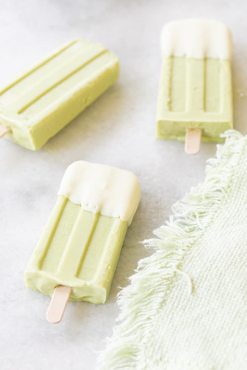 Three green avocado popsicles dipped in white chocolate on a marble table with green towel. 