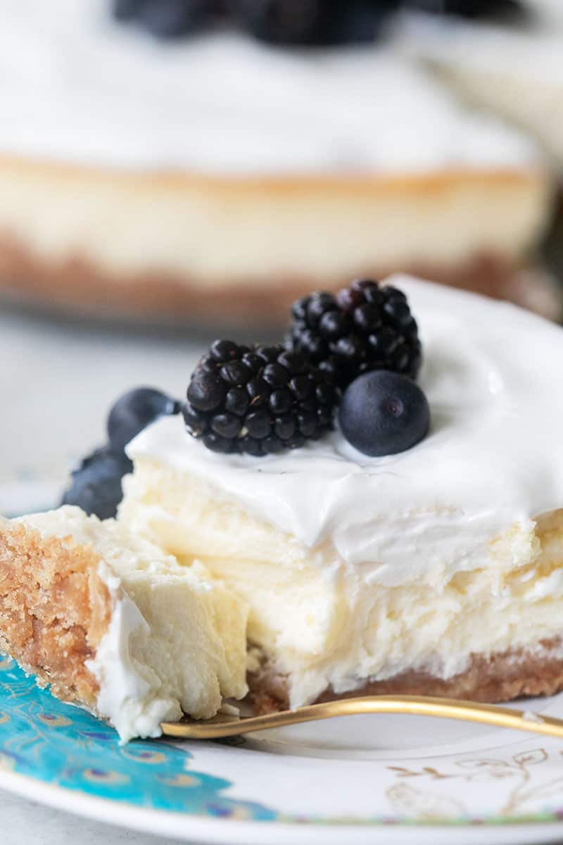 Slice of cheesecake with sour cream topping and blackberries on top. Served with a gold spoon. 