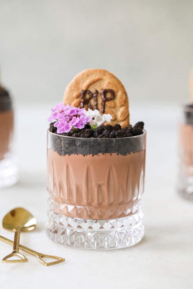 A glass of chocolate pudding with Oreo crumbs on top with a tombstone cookie and flowers.