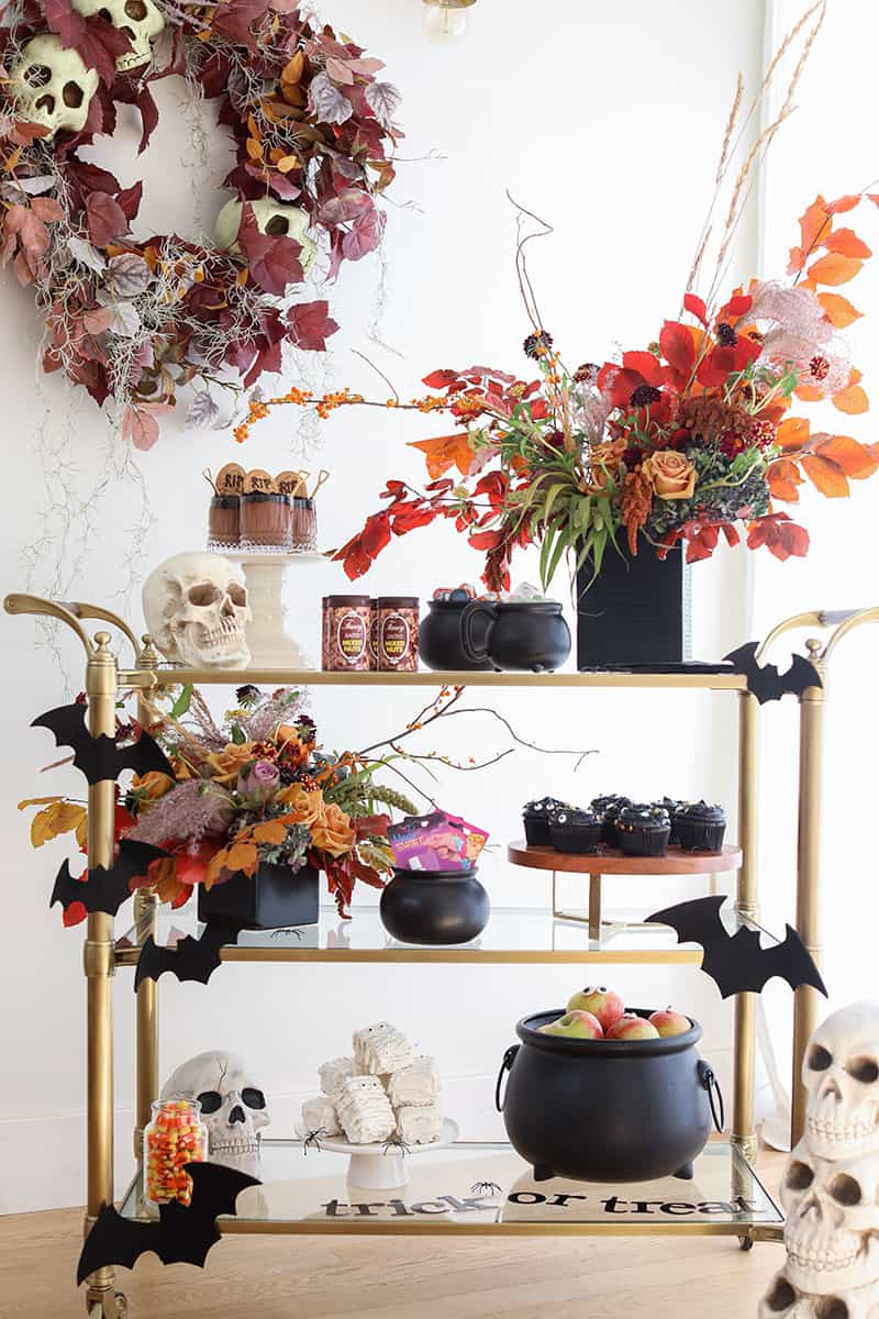 Halloween party bar cart filled with tricks and treats like cupcakes, mummy rice krispies treats, skulls, bats and flowers.