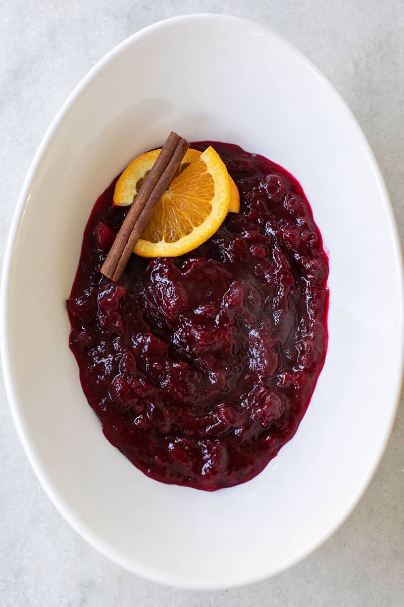 Thanksgiving cranberry sauce recipe served in a white oval bowl with orange slices and a cinnamon stick. 