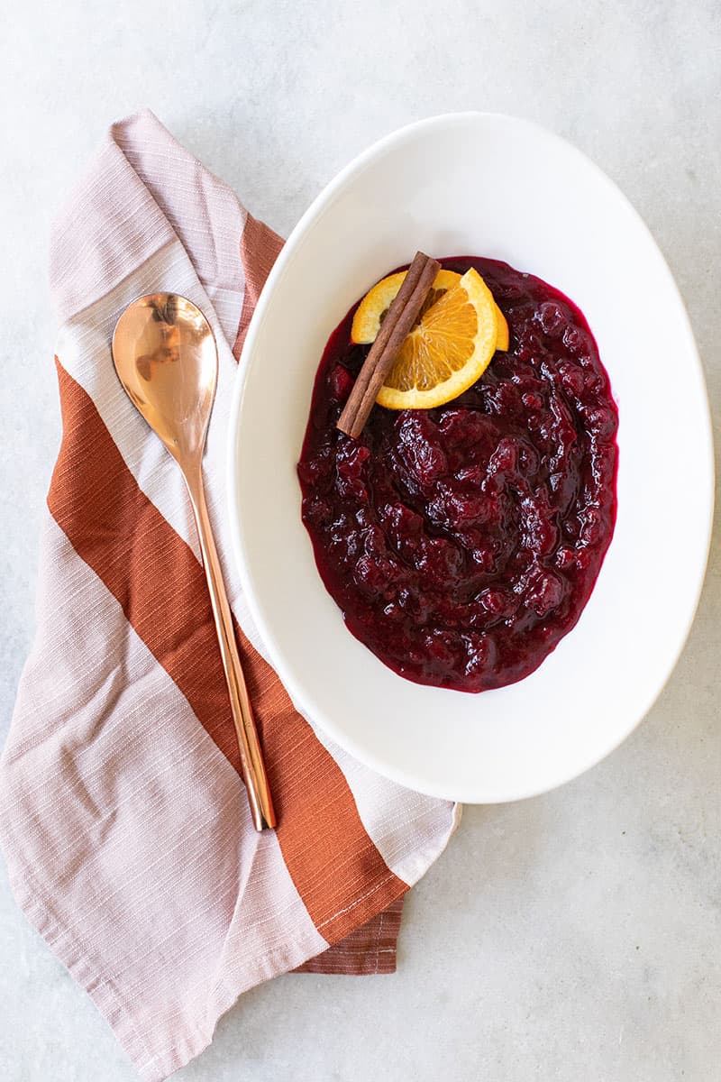 Thanksgiving cranberry sauce with oranges and cinnamon sticks. Served in an oval bowl with a copper spoon and striped towel. 