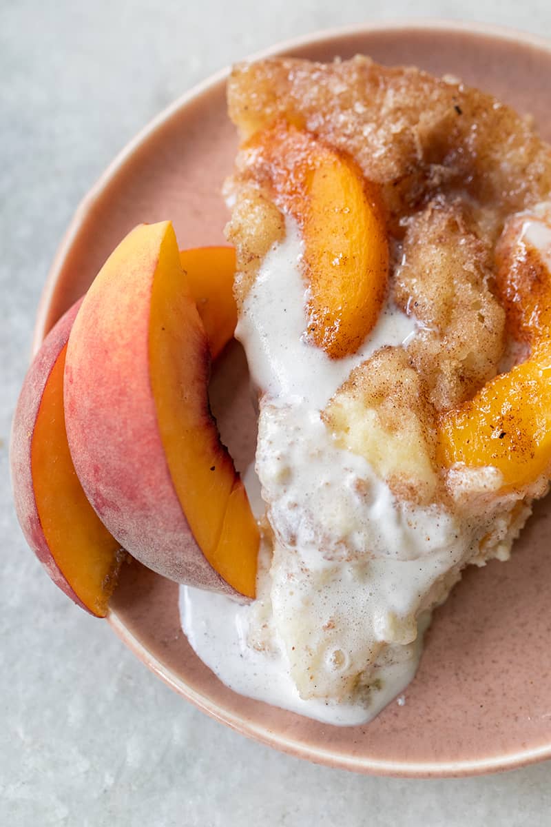 Warm slice of peach cake with melted vanilla ice cream and fresh peaches. Served on a pink plate.