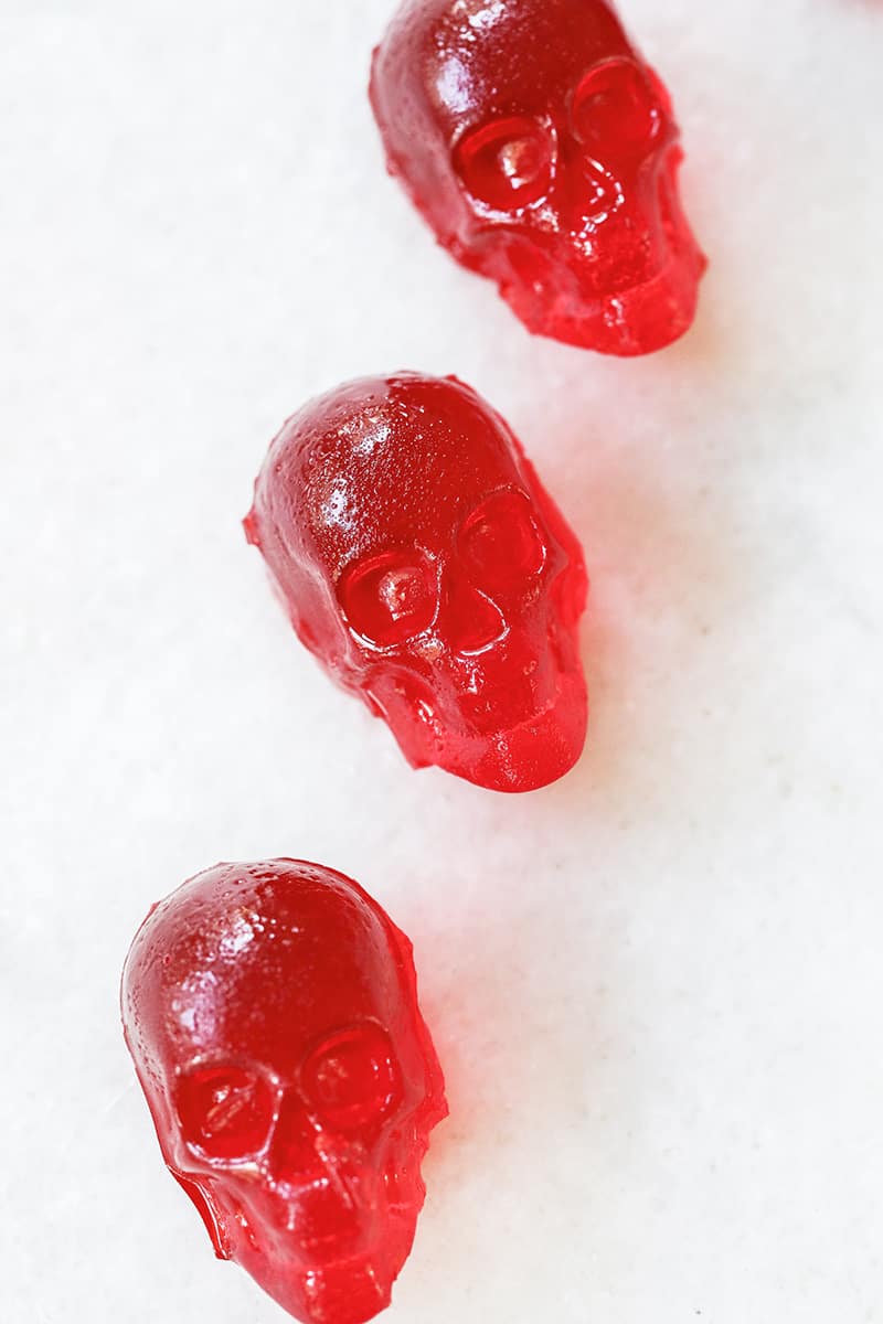 Bright red cherry jello shots shaped like skulls on a marble table.