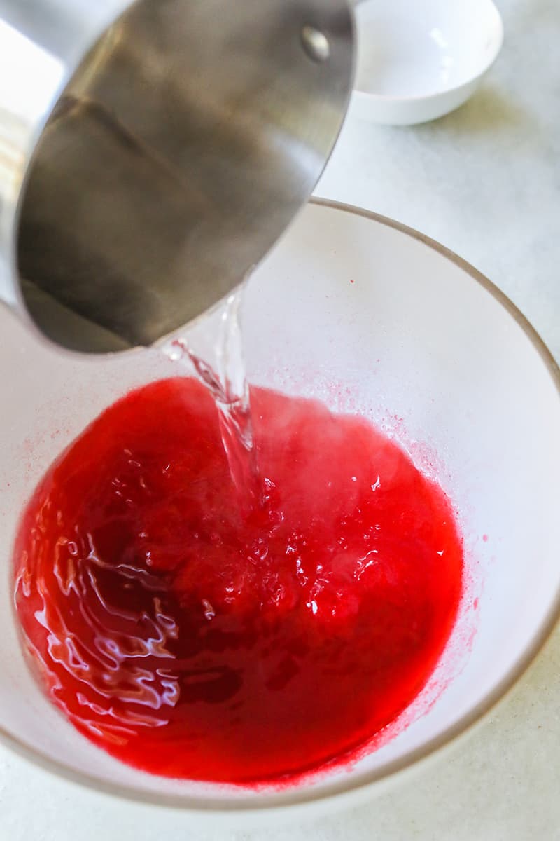 Boiling water being poured into a white bowl with red jello, vodka and gelatin.