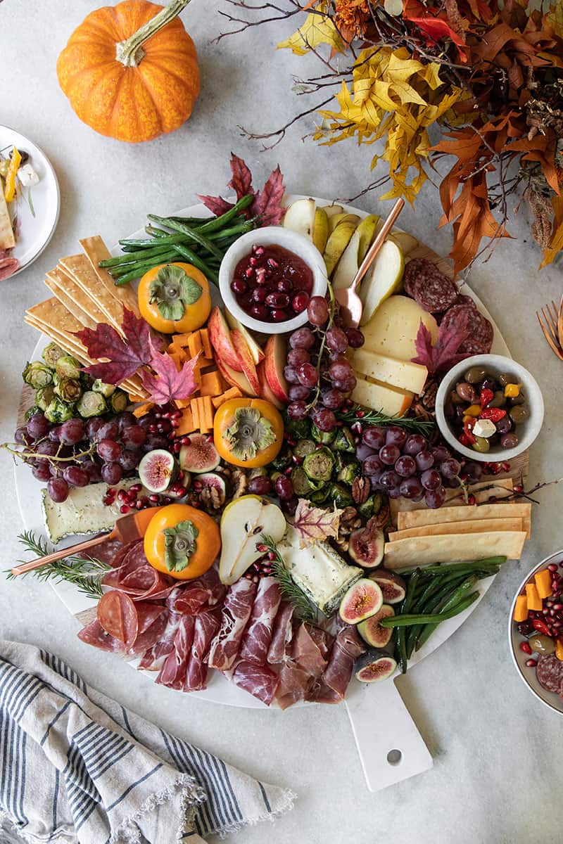 A Thanksgiving appetizer cheeseboard with fruit, cheese, meat, crackers, olives and more. On a marble table with fall leaves.